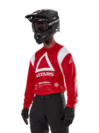Alpinestars Techdura Jersey, Motocross-Trikot, leuchtendes Rot und Weiß, mit markantem weißem Dreiecksdesign und ASTARS-Branding, getragen mit schwarzem Helm und Handschuhen, strapazierfähige und feuchtigkeitsableitende Offroad-Rennausrüstung