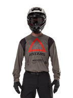 Motocross-Fahrer im Alpinestars Techdura Jersey in Falcon Brown, mit einem markanten roten Dreieck und „ASTARS“-Branding auf der Brust, kombiniert mit schwarzer Hose, schwarzem Helm mit Brille und schwarzen Handschuhen, komplette Enduro-Rennausrüstung.