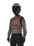 Alpinestars Techdura Jersey, Motocross-Offroad-Trikot, Falcon Brown mit rotem Dreieck und schwarzem Branding, getragen mit schwarzer Hose, schwarz-weißem Helm und schwarzen Handschuhen