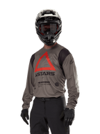 Alpinestars Techdura Jersey, Motocross-Offroad-Trikot, Falcon Brown mit rotem Dreieck und schwarzem Branding, getragen mit schwarzer Hose, schwarz-weißem Helm und schwarzen Handschuhen