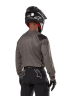 Motocross-Fahrer von hinten im Alpinestars Techdura Jersey in Falcon Brown, kombiniert mit einer schwarzen Offroad-Hose und einem schwarzen Helm, modernes Grafikdesign und verlängerte Rückseite für Enduro-Fahrten