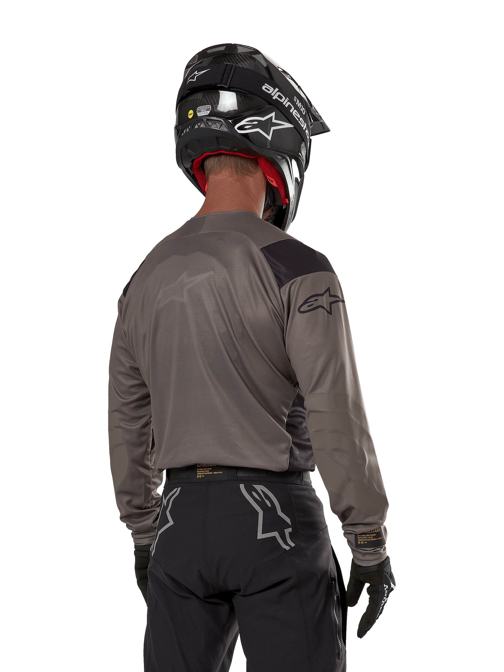 Motocross-Fahrer von hinten im Alpinestars Techdura Jersey in Falcon Brown, kombiniert mit einer schwarzen Offroad-Hose und einem schwarzen Helm, modernes Grafikdesign und verlängerte Rückseite für Enduro-Fahrten