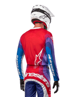 Motocross-Fahrer von hinten im Alpinestars 2024 Techstar Pneuma Jersey in Blue Mars Red White, mit einer roten Rückseite und weißem Stern-Logo, blau-violetten Ärmeln mit weißem Branding, kombiniert mit passender Hose und einem weißen Helm für den Offroad-Rennsport.