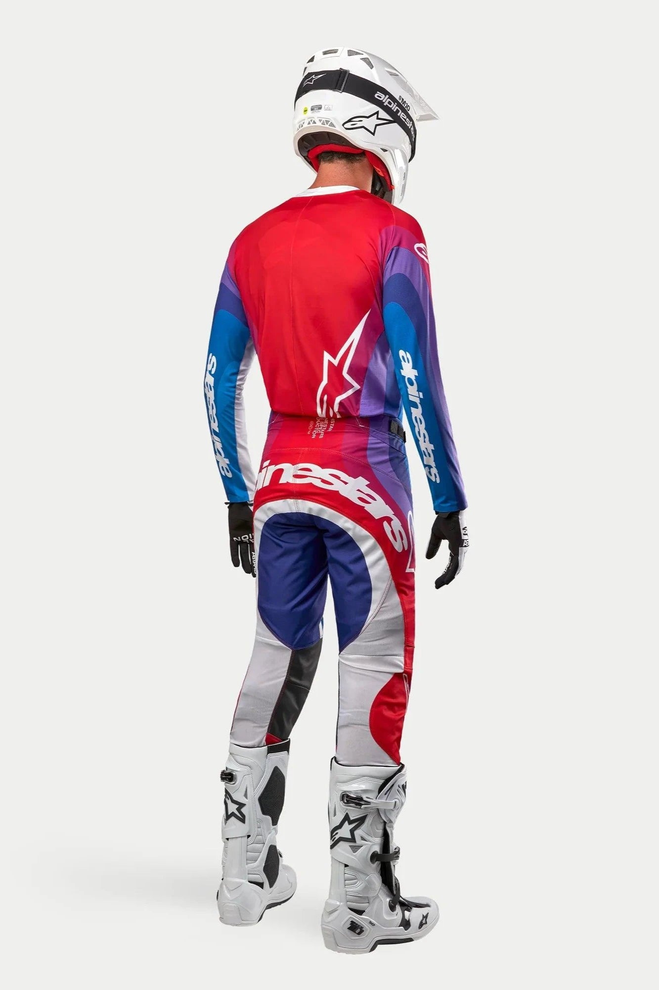 Motocross-Fahrer von hinten in Alpinestars 2024 Techstar Pneuma Trikot und Hose, Farbvariante Blu Mars Red White mit rot-blau-lila Farbverläufen und weißem Branding, kombiniert mit weißen Stiefeln und einem weißen Helm, koordinierte Offroad-Rennausrüstung.