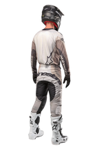 Motocross-Fahrer in der Rückansicht im 2024 Techstar Pneuma Trikot in Dark Sand Iron Dust Gray, passende Motocross-Hose mit markantem Branding, schwarzer Helm und weiße Rennstiefel, abgestimmte professionelle Offroad-Ausrüstung.