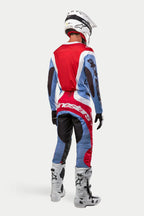 Motocross-Fahrer in kompletter Rennausrüstung von hinten, 2024 Techstar Ocuri Trikot und Hose in Hellblau, Marsrot und Weiß, weißer Alpinestars-Helm mit schwarzen Grafiken, weiß-graue Offroad-Stiefel, schwarze Handschuhe, abgestimmtes Outfit für Motocross-Rennen