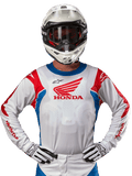 Honda Racer Iconic Trikot, weißes Motocross-Trikot mit rotem Honda-Logo und leuchtend blauen und roten Akzenten, getragen von einem Fahrer mit weißem Helm und Handschuhen, Performance-Ausrüstung für Offroad- und Motocross-Rennsport