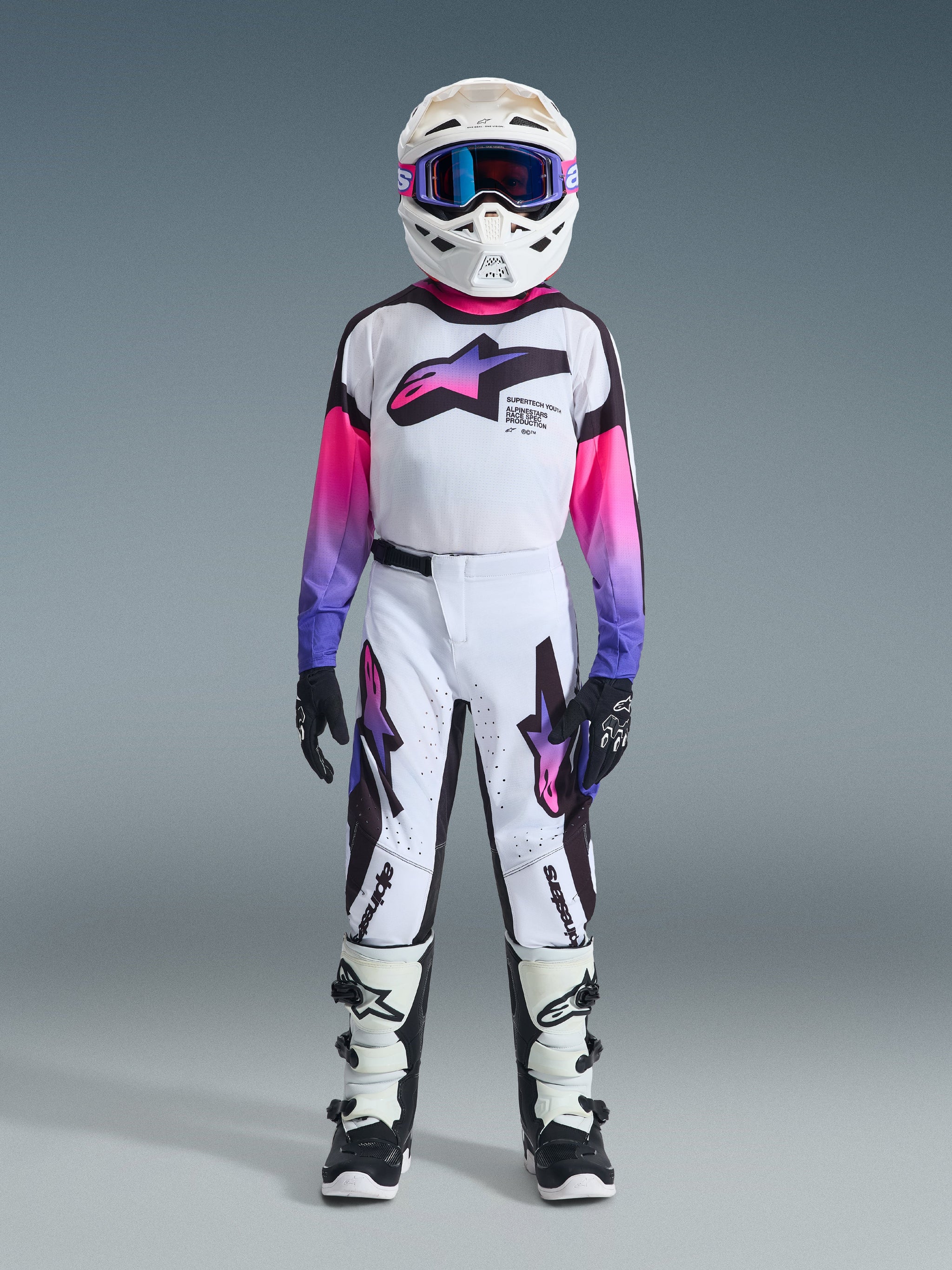 Youth motocross rider wearing complete racing gear, 2026 Youth Supertech Vista Jersey in white with pink and purple gradient sleeves, matching white and black pants with purple accents, white helmet with purple goggles, black and white boots, black gloves, coordinated high-performance outfit for off-road racing