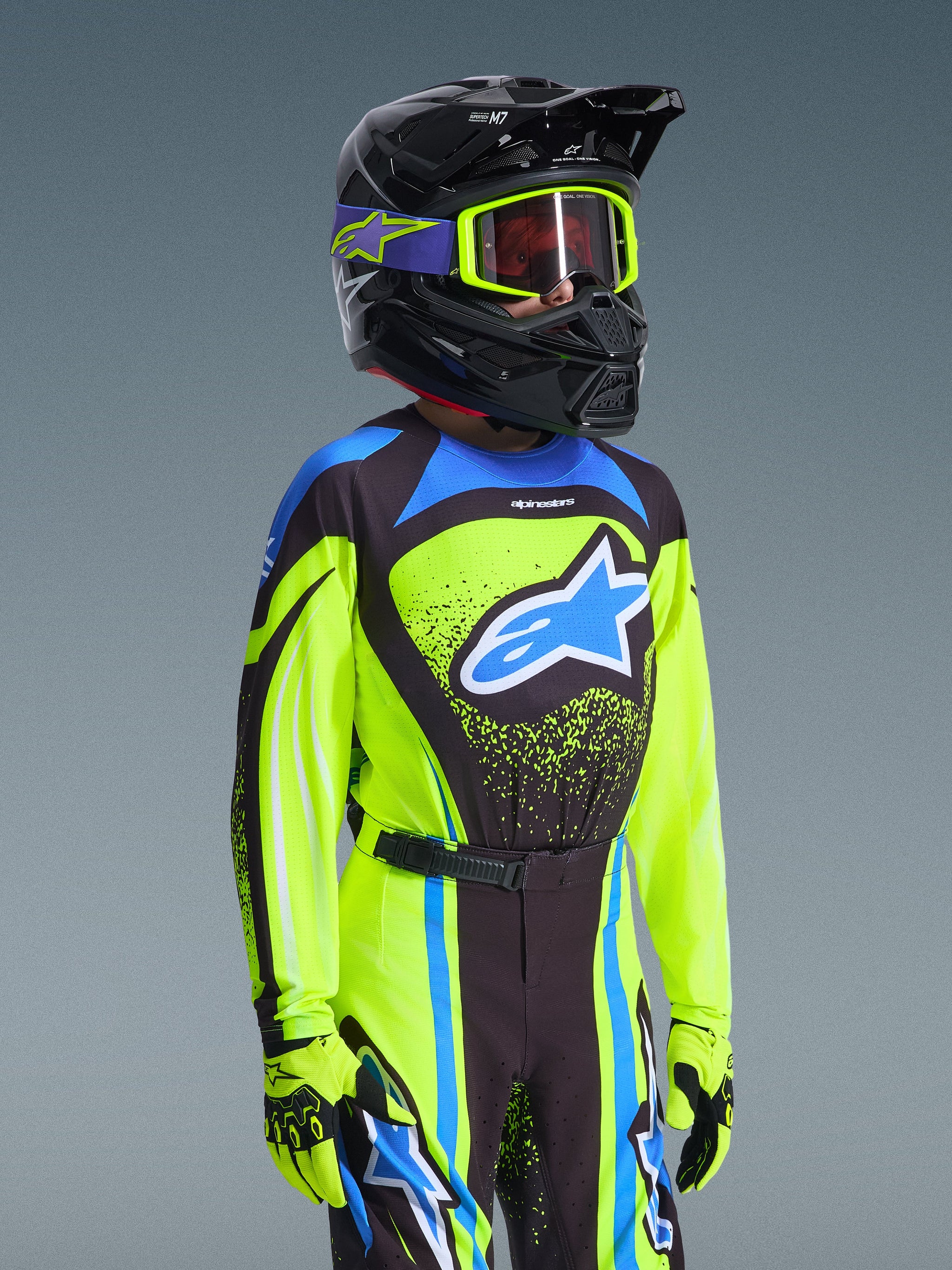 Jugend-Motocross-Fahrer mit Alpinestars 2026 Youth Supertech Nomur Jersey in Gelb Fluo und Dunkelblau, blaues Astars-Logo, passende Rennhose und Handschuhe, schwarzer Helm mit fluogelber Brille, professionelle Offroad-Motocross-Ausrüstung.