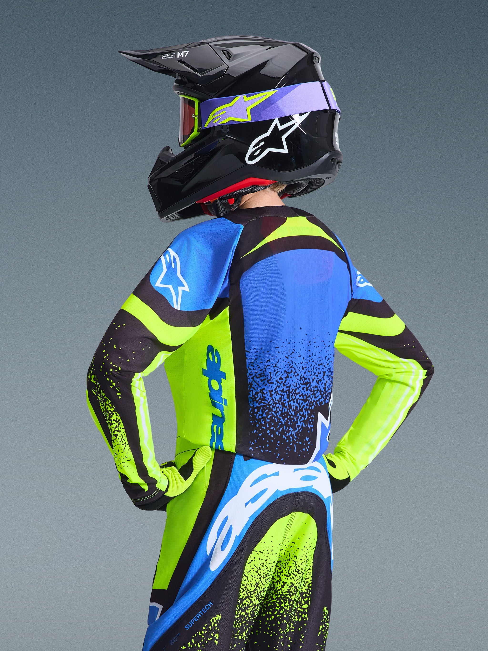 Jugend-Motocross-Fahrer von hinten im Alpinestars 2026 Youth Supertech Nomur Jersey, Dunkelblau und Gelb Fluo mit blauen Akzenten, kombiniert mit passendem schwarzem Helm und Hose, professionelle Offroad-Rennausrüstung.