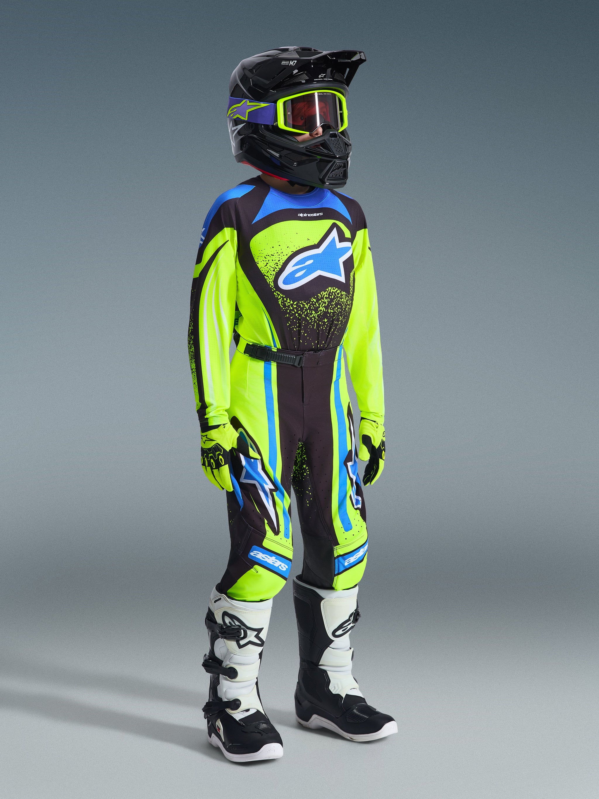 Jugend-Motocross-Fahrer in kompletter Rennausrüstung, 2026 Youth Supertech Nomur Jersey in Gelb Fluo und Dunkelblau, passende Hose mit blauen Akzenten, schwarzer Helm mit neongelber Brille, weiß-schwarze Stiefel, koordiniertes Outfit für den Offroad-Rennsport