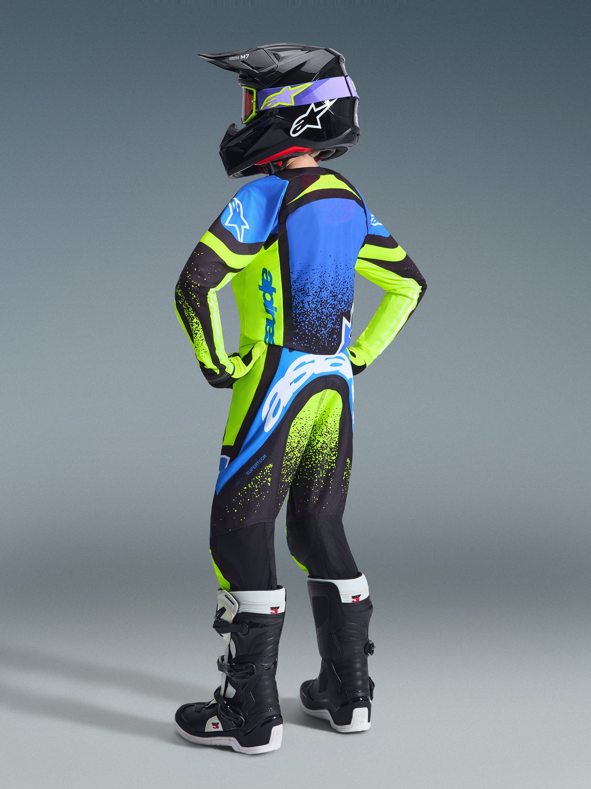 Alpinestars 2026 Youth Supertech Nomur Jersey und Hose, Jugend-Motocross-Fahrer von hinten in voller Rennausrüstung, leuchtendes Gelb-Fluo und Dunkelblau mit UCLA-Blau-Akzenten, passender schwarzer Helm und Stiefel, abgestimmtes Profi-Offroad-Outfit