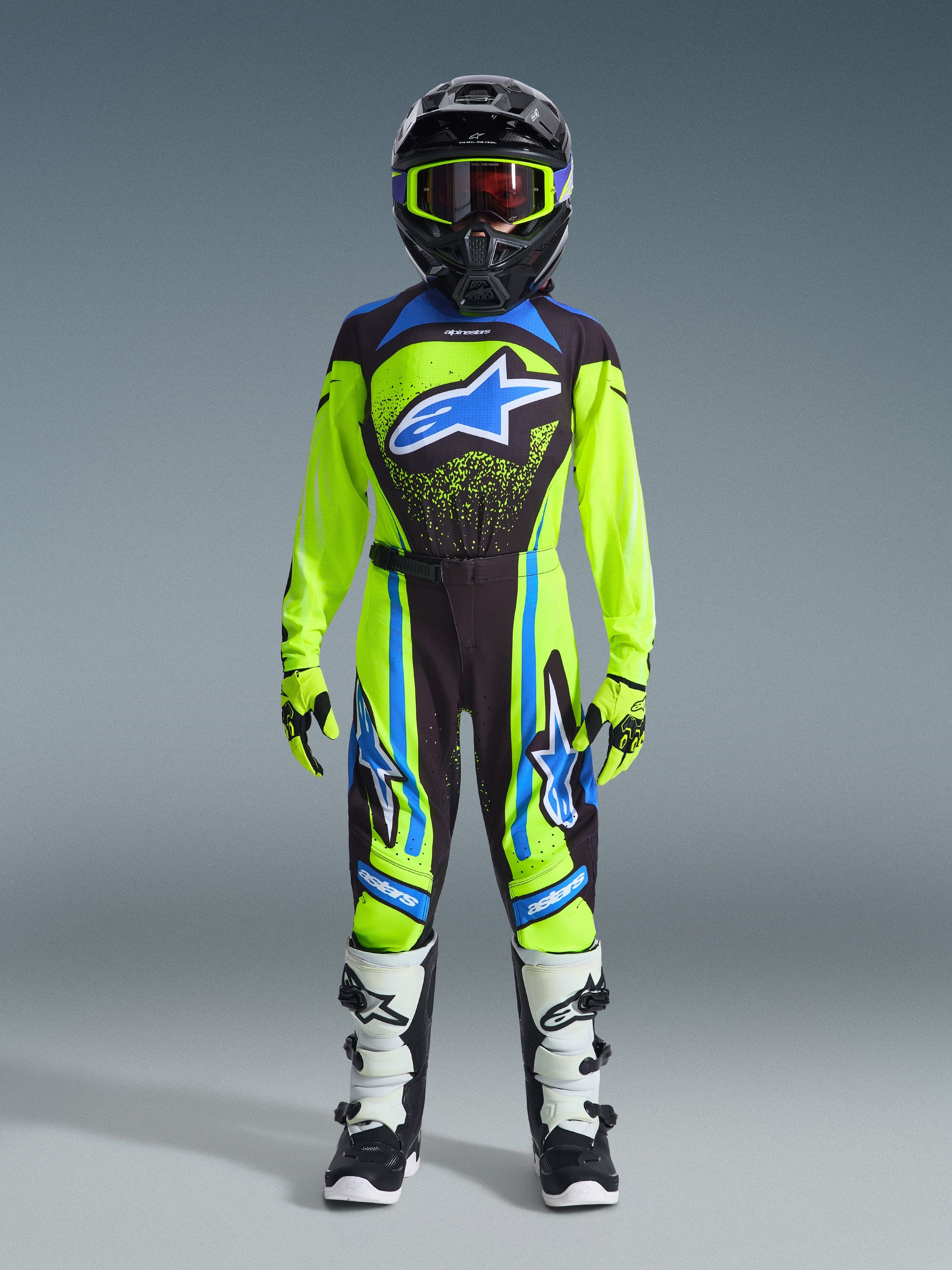 Jugend-Motocross-Fahrer in kompletter Rennausrüstung, 2026 Jugend Supertech Nomur Trikot in Neon-Gelb und Dunkelblau mit blauem Alpinestars-Logo, passende Hose, weiß-schwarze Stiefel, neon-gelbe Handschuhe und schwarzer Helm mit Schutzbrille, professionelles Offroad-Rennoutfit.