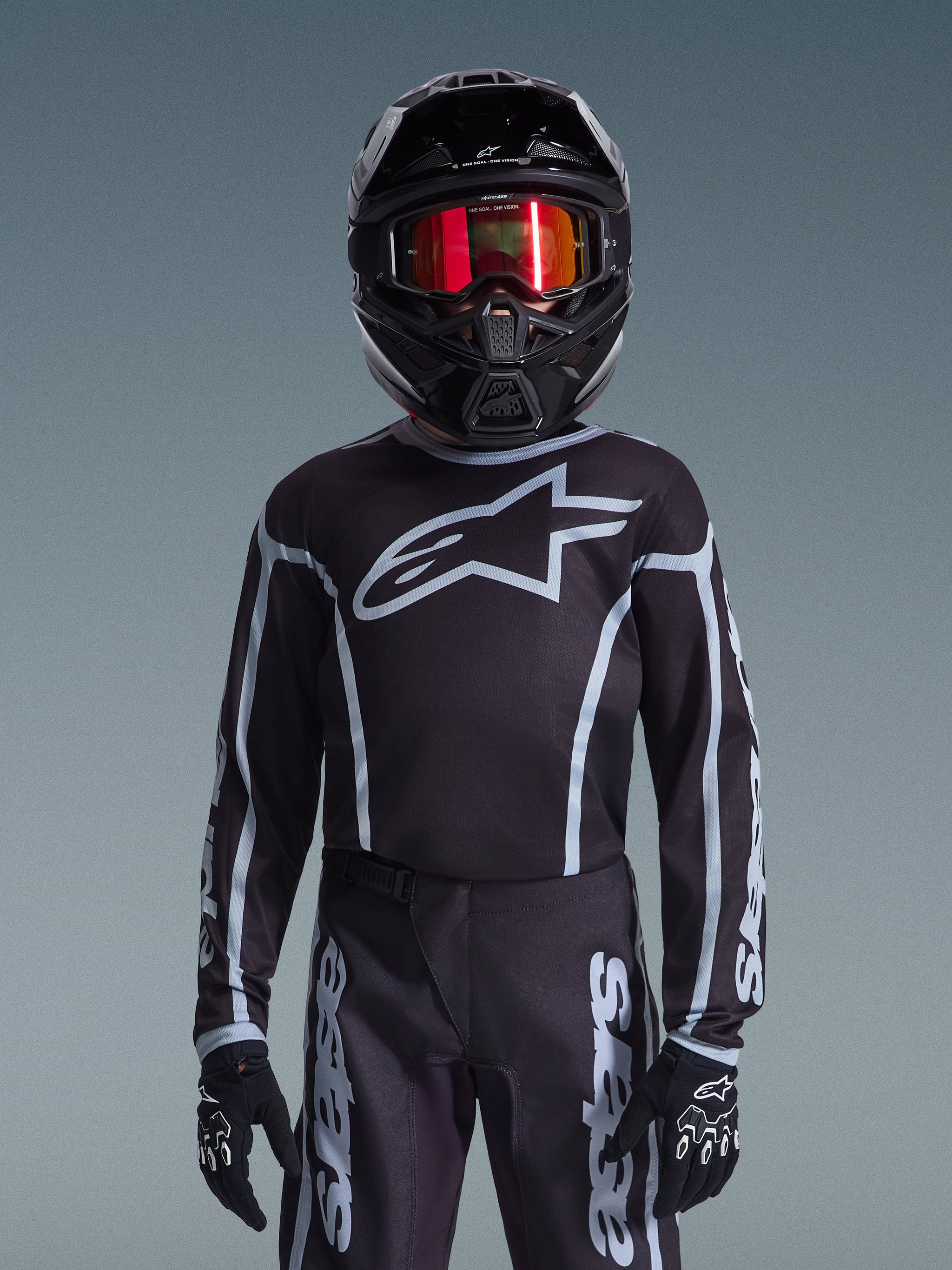 Jugend-Motocross-Fahrer im Alpinestars 2026 Youth Fluid Apex Trikot in Schwarz und Grau, passende Hose, schwarzer Helm mit rot reflektierender Brille und schwarzen Handschuhen, koordinierte Offroad-Rennausrüstung