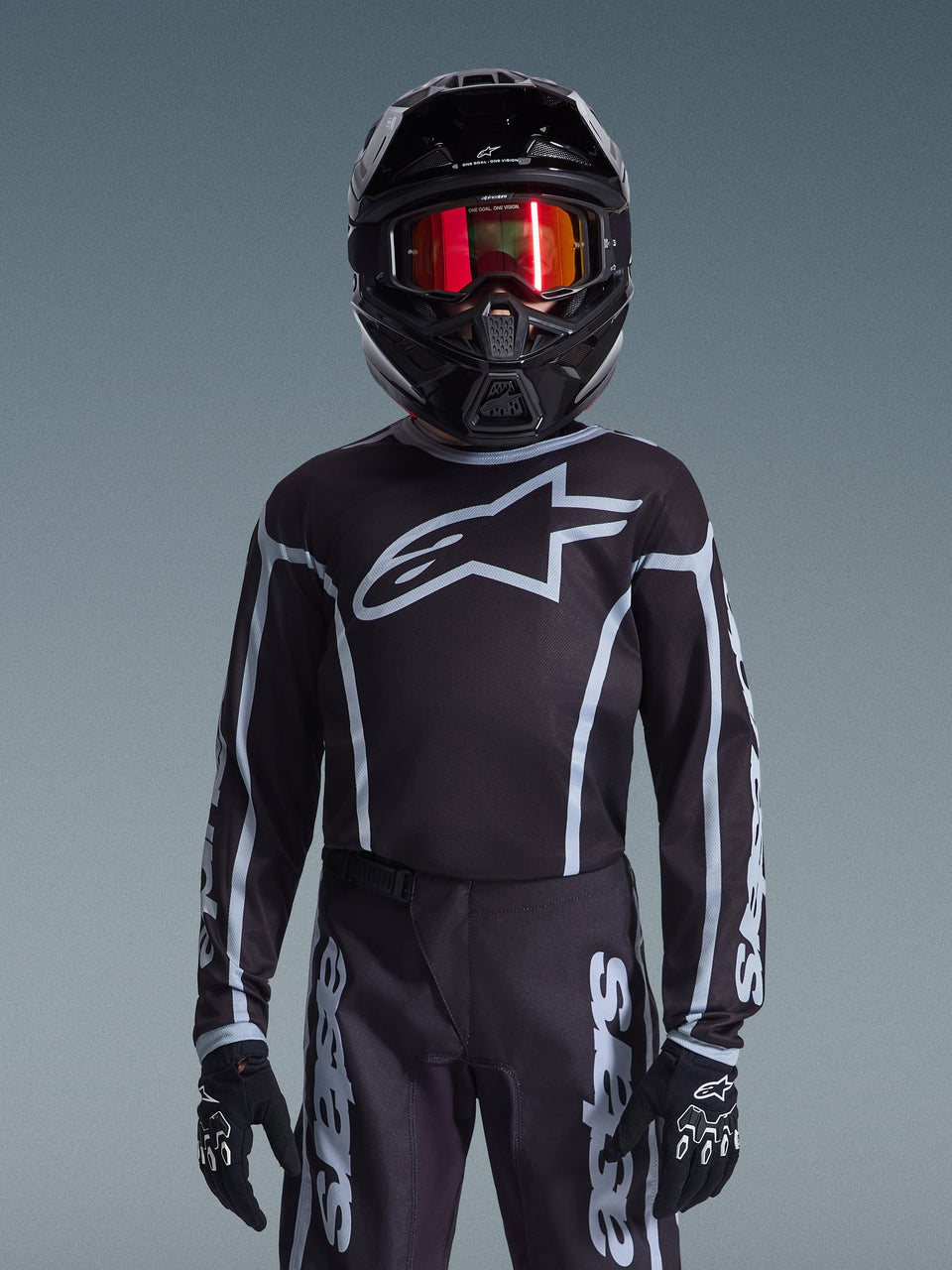 Jugend-Motocross-Fahrer im Alpinestars 2026 Youth Fluid Apex Trikot in Schwarz und Grau, passende Hose, schwarzer Helm mit rot reflektierender Brille und schwarzen Handschuhen, koordinierte Offroad-Rennausrüstung