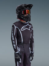 Alpinestars 2026 Youth Fluid Apex Trikot in Schwarz und Grau, getragen von einem jungen Fahrer mit passendem schwarzem Helm und getönter Brille, auffälliges Alpinestars-Branding auf Brust und Ärmeln, Teil eines abgestimmten Motocross-Ausrüstungssets.