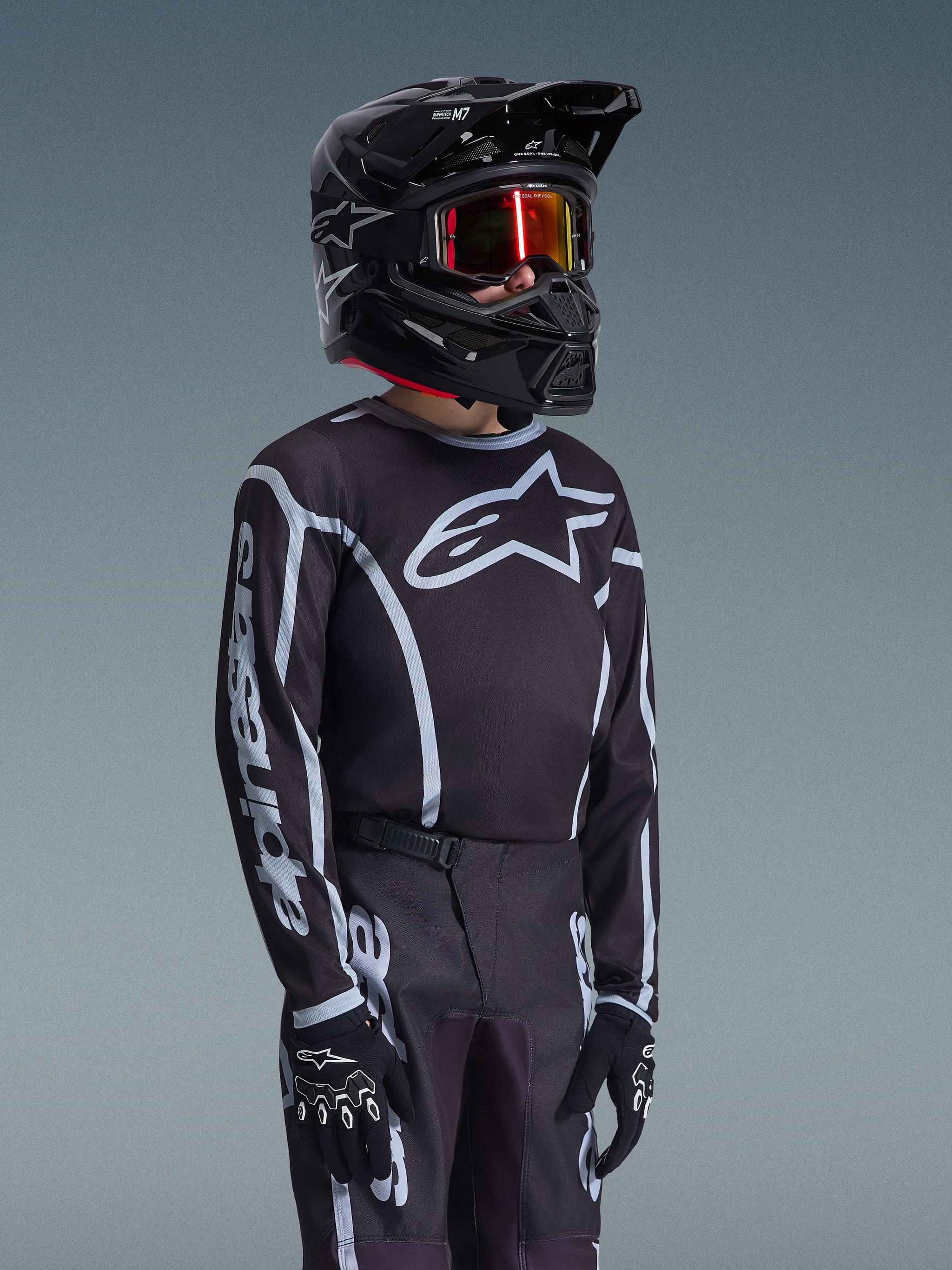 Alpinestars 2026 Youth Fluid Apex Trikot in Schwarz und Grau, getragen von einem jungen Fahrer mit passendem schwarzem Helm und getönter Brille, auffälliges Alpinestars-Branding auf Brust und Ärmeln, Teil eines abgestimmten Motocross-Ausrüstungssets.