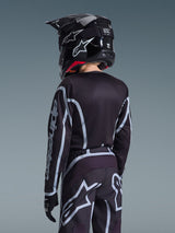 Rückansicht eines jugendlichen Motocross-Fahrers im Alpinestars 2026 Youth Fluid Apex Trikot und der passenden Hose in Schwarz und Grau, schwarzer Helm mit weißem Branding, schwarze Handschuhe, sportliche Offroad-Rennausrüstung für junge Fahrer.