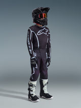 Jugend-Motocross-Fahrer im 2026 Youth Fluid Apex Trikot in Schwarz-Grau, passende schwarz-graue Hose mit weißen Grafiken, schwarzer Helm mit rot getönter Brille, schwarze Handschuhe und weiß-schwarze Motocross-Stiefel, komplette Offroad-Rennausrüstung.