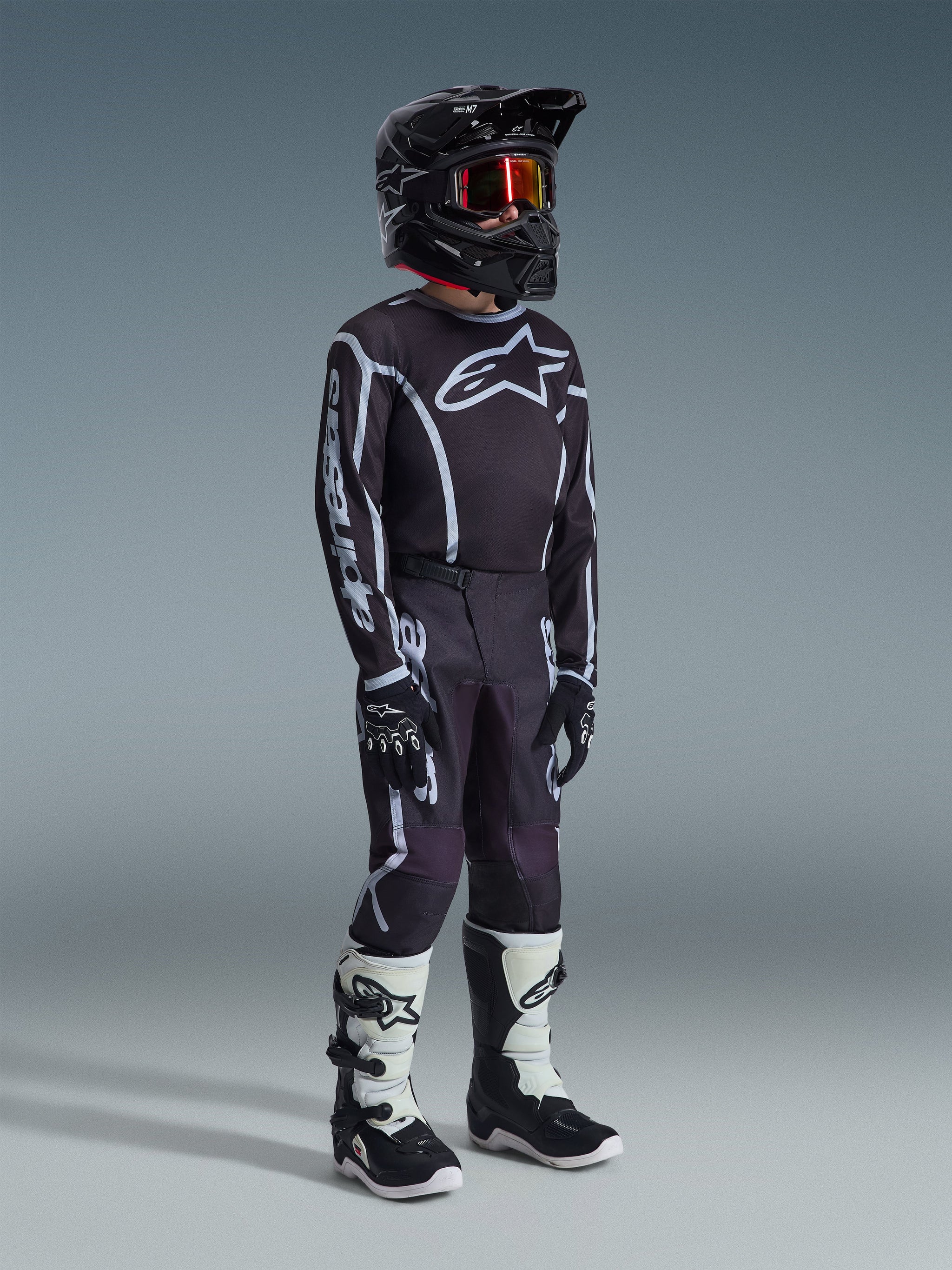 Jugend-Motocross-Fahrer im 2026 Youth Fluid Apex Trikot in Schwarz-Grau, passende schwarz-graue Hose mit weißen Grafiken, schwarzer Helm mit rot getönter Brille, schwarze Handschuhe und weiß-schwarze Motocross-Stiefel, komplette Offroad-Rennausrüstung.