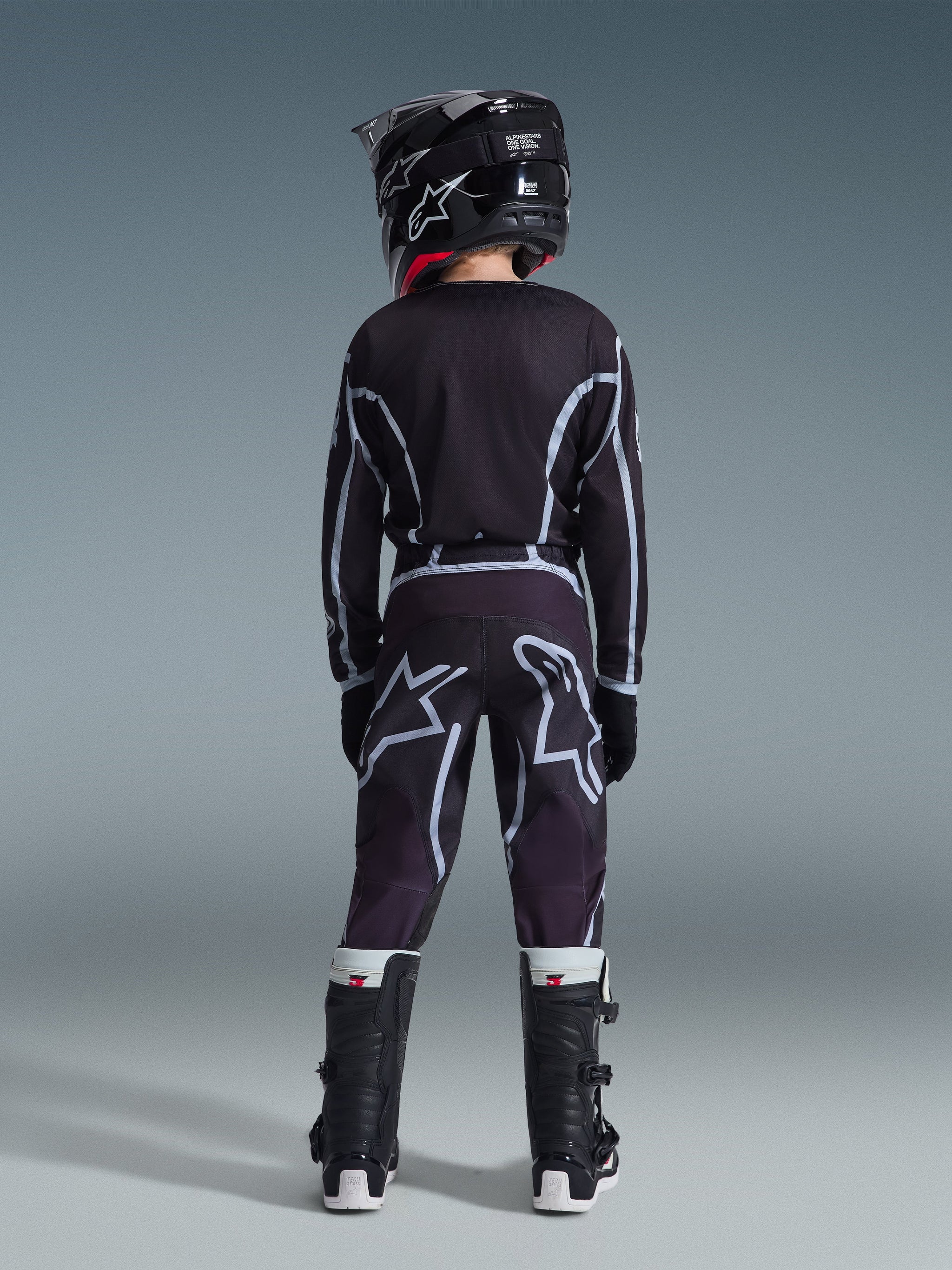 Jugendlicher Motocross-Fahrer von hinten gesehen im 2026 Youth Fluid Apex Trikot in Schwarz und Grau, passende Motocross-Hose mit großen weißen Logo-Grafiken, schwarzer Helm und schwarz-weiße Tech Series Stiefel, komplette Performance-Ausrüstung für das Offroad-Fahren.