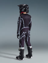Alpinestars 2026 Youth Fluid Apex Trikot, Motocross-Trikot, Schwarz und Grau, getragen mit passender Youth Fluid Grid Hose, schwarzem Helm und schwarz-weißen Stiefeln, Rückansicht der kompletten Offroad-Rennausrüstung für junge Fahrer