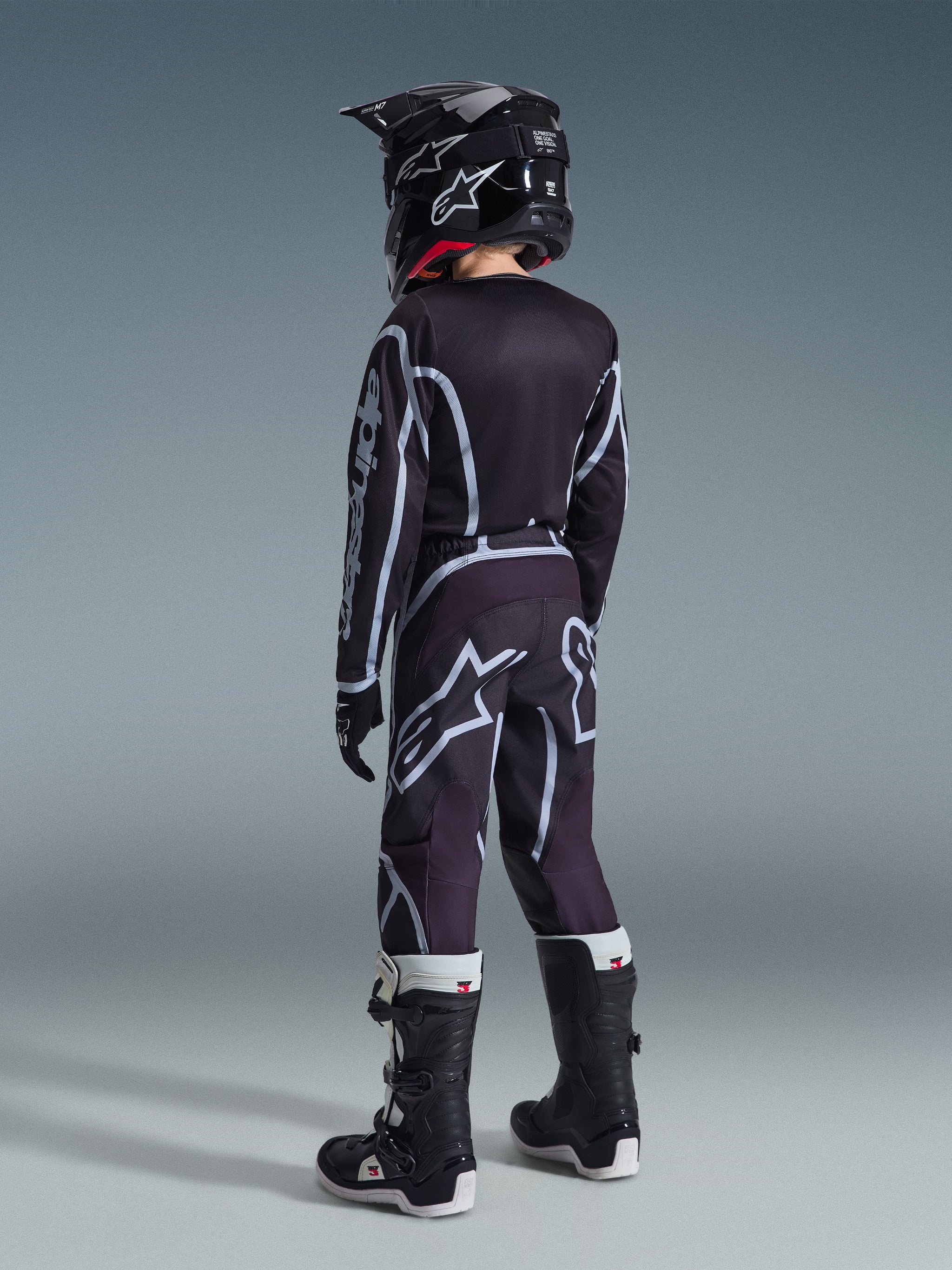 Alpinestars 2026 Youth Fluid Apex Trikot, Motocross-Trikot, Schwarz und Grau, getragen mit passender Youth Fluid Grid Hose, schwarzem Helm und schwarz-weißen Stiefeln, Rückansicht der kompletten Offroad-Rennausrüstung für junge Fahrer
