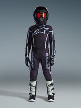 Junger Motocross-Fahrer im Alpinestars 2026 Youth Fluid Apex Trikot in Schwarz und Grau, kombiniert mit passender Hose, schwarzem Helm mit rot irisierender Brille, schwarz-weißen Handschuhen und Motocross-Stiefeln, komplette Ausrüstung für den Jugend-Offroad-Rennsport