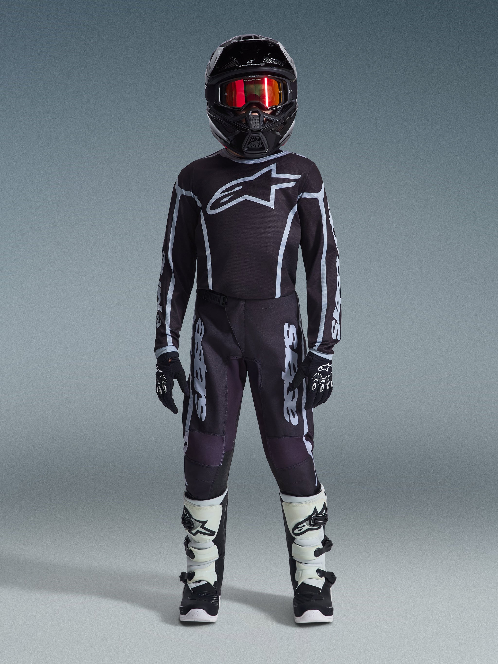 Junger Motocross-Fahrer im Alpinestars 2026 Youth Fluid Apex Trikot in Schwarz und Grau, kombiniert mit passender Hose, schwarzem Helm mit rot irisierender Brille, schwarz-weißen Handschuhen und Motocross-Stiefeln, komplette Ausrüstung für den Jugend-Offroad-Rennsport