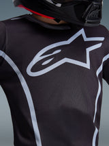 Alpinestars 2026 Youth Fluid Apex Trikot, Nahaufnahme des Brustdesigns, Schwarz und Grau, mit einem markanten hellgrauen Stern-Logo und sportlichen Paspeln, getragen mit einem schwarzen Helm, Performance-Motocross-Ausrüstung für junge Fahrer