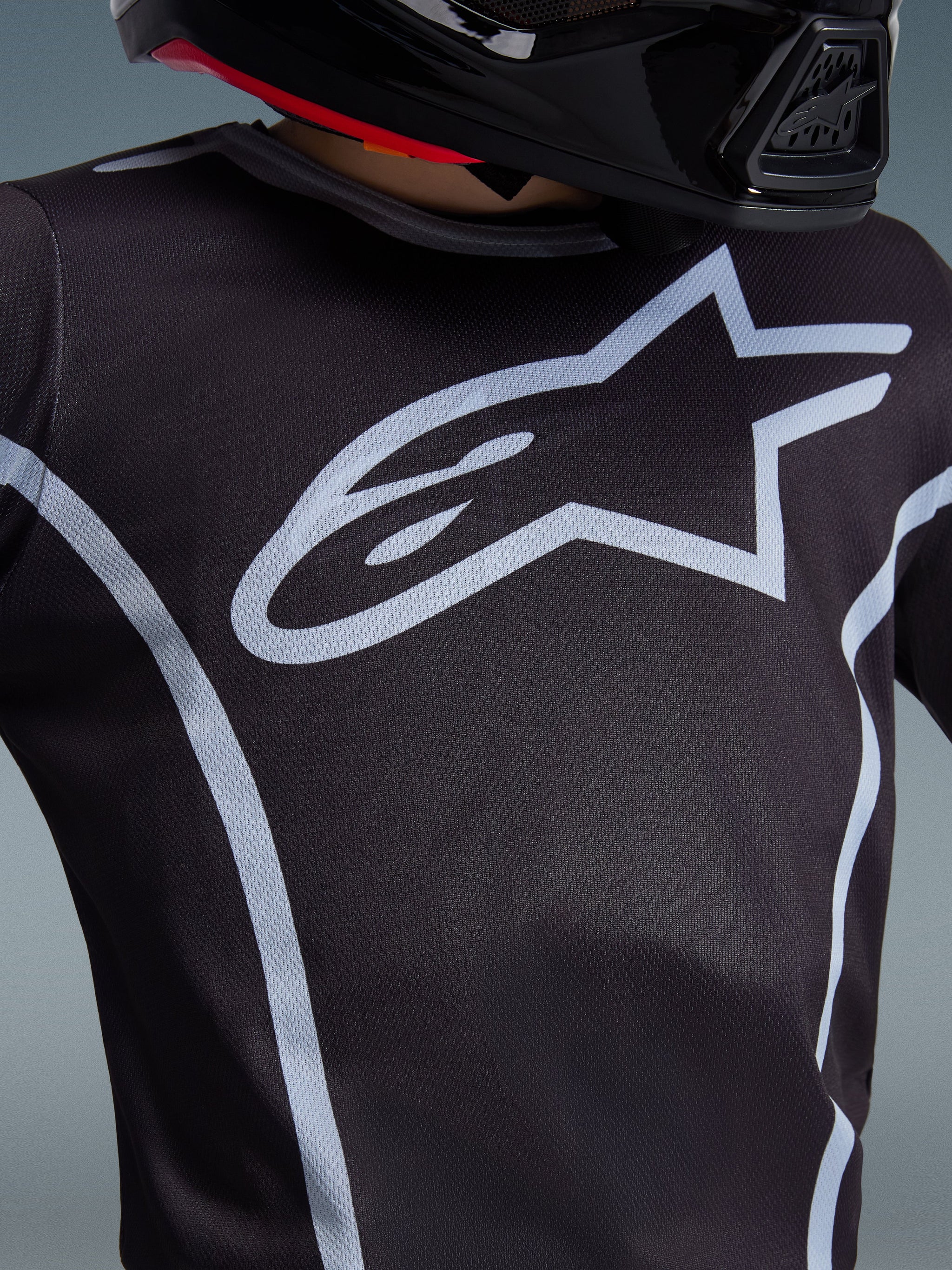 Alpinestars 2026 Youth Fluid Apex Trikot, Nahaufnahme des Brustdesigns, Schwarz und Grau, mit einem markanten hellgrauen Stern-Logo und sportlichen Paspeln, getragen mit einem schwarzen Helm, Performance-Motocross-Ausrüstung für junge Fahrer