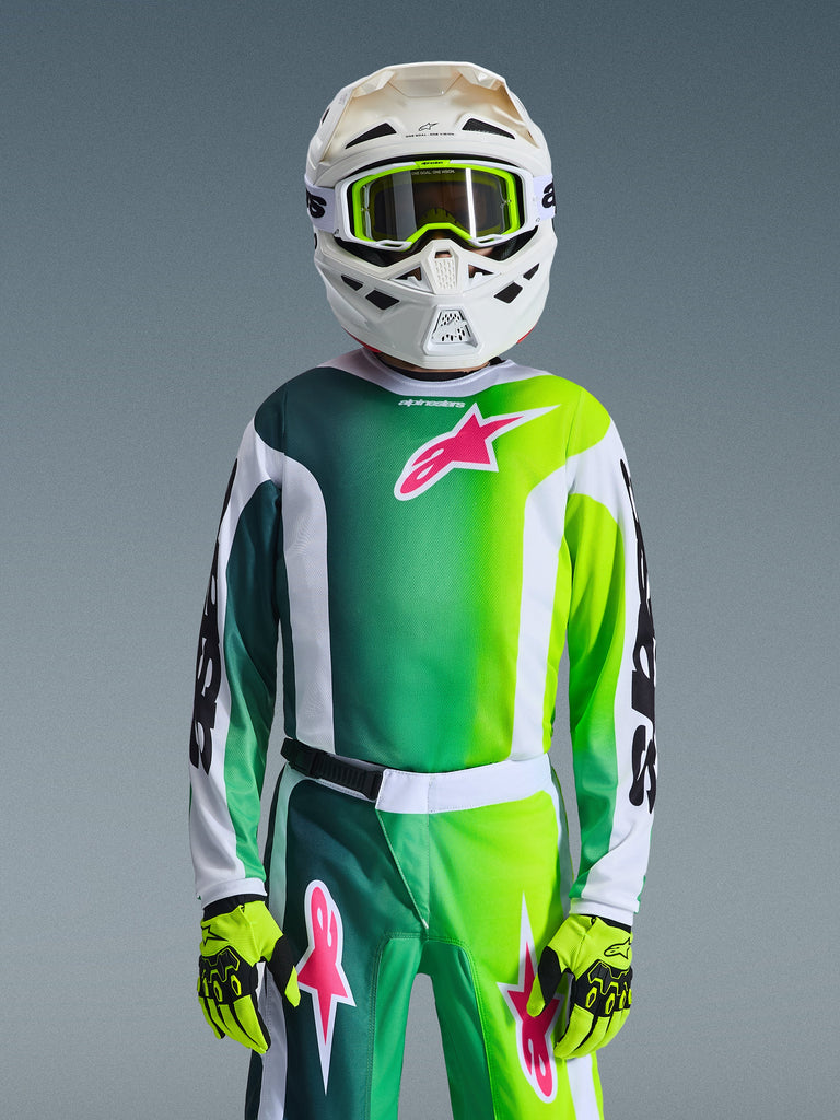 Jugend-Motocross-Fahrer in kompletter Rennausrüstung, grün-weiß-schwarzes Jugend-Fluid Portl Trikot 2026 mit pinkem Logo-Akzent, passende Hose und Handschuhe, weißer Motocross-Helm mit limonengrüner Brille, koordiniertes Outfit für den Offroad-Rennsport