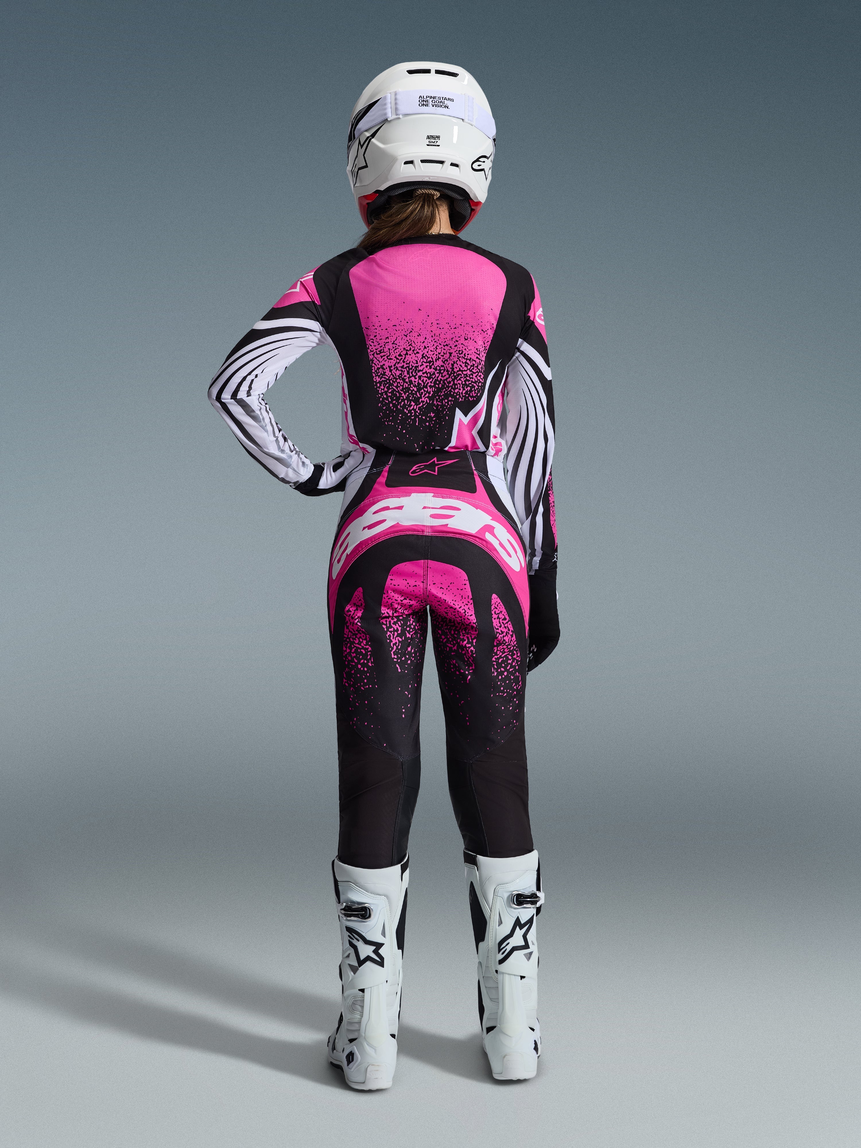 Alpinestars 2026 Stella Techstar Nomur Gear Set, Motocross-Fahrerin in der Rückansicht, pinkfarbenes und schwarzes Jersey mit weiß gestreiften Ärmeln und Sprenkelmuster, passende Hose mit weißem Alpinestars-Logo, weißer Helm und weiße Offroad-Stiefel