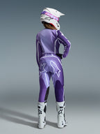 Alpinestars 2026 Stella Fluid Apex Gear Set, Damen-Motocross-Ausrüstung, Lila und Lavendel mit weißen Akzenten, Fahrerin von hinten in passendem Trikot und Hose, weißer Helm mit pinker Brille, weiße Stiefel, koordiniertes sportliches Rennoutfit