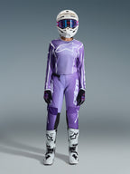 Motocross-Fahrerin im Alpinestars 2026 Stella Fluid Apex Gear Set, lila Jersey mit weißem Logo, passende lila Hose mit weißem Branding, weißer Helm mit pinker Brille, weiß-schwarze Stiefel, abgestimmtes Damen-Motocross-Outfit für Offroad-Rennen