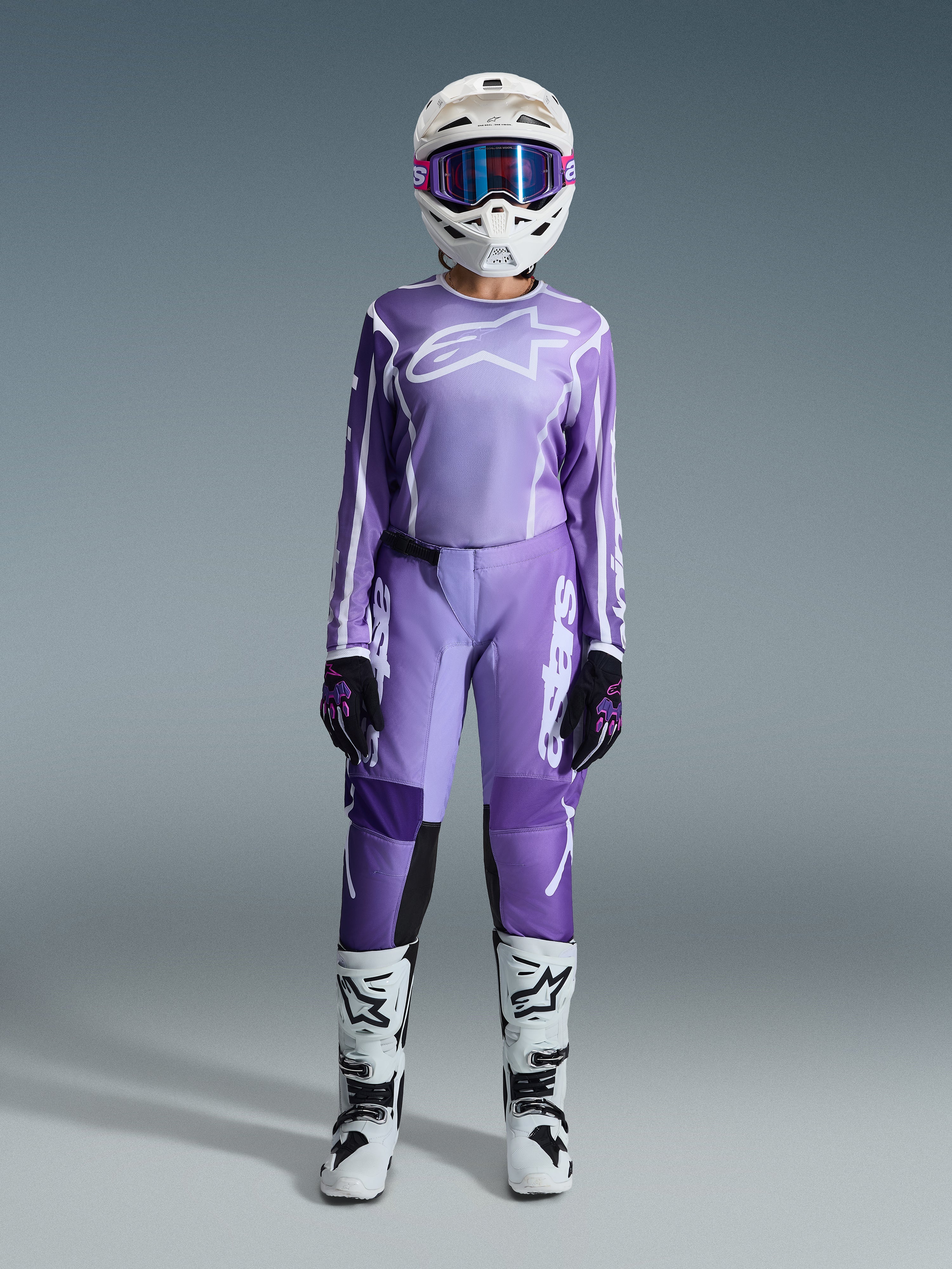 Motocross-Fahrerin im Alpinestars 2026 Stella Fluid Apex Gear Set, lila Jersey mit weißem Logo, passende lila Hose mit weißem Branding, weißer Helm mit pinker Brille, weiß-schwarze Stiefel, abgestimmtes Damen-Motocross-Outfit für Offroad-Rennen