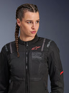Alpinestars Stella ST-2 Air Jacke, Motorradjacke für Damen in Schwarz, mit Mesh-Einsätzen für Luftzirkulation und „Alpinestars Exploration Equipment“-Schriftzug mit Koordinaten, rotes Logo-Detail auf der Brust, getragen von einem Model mit geflochtenem Haar.