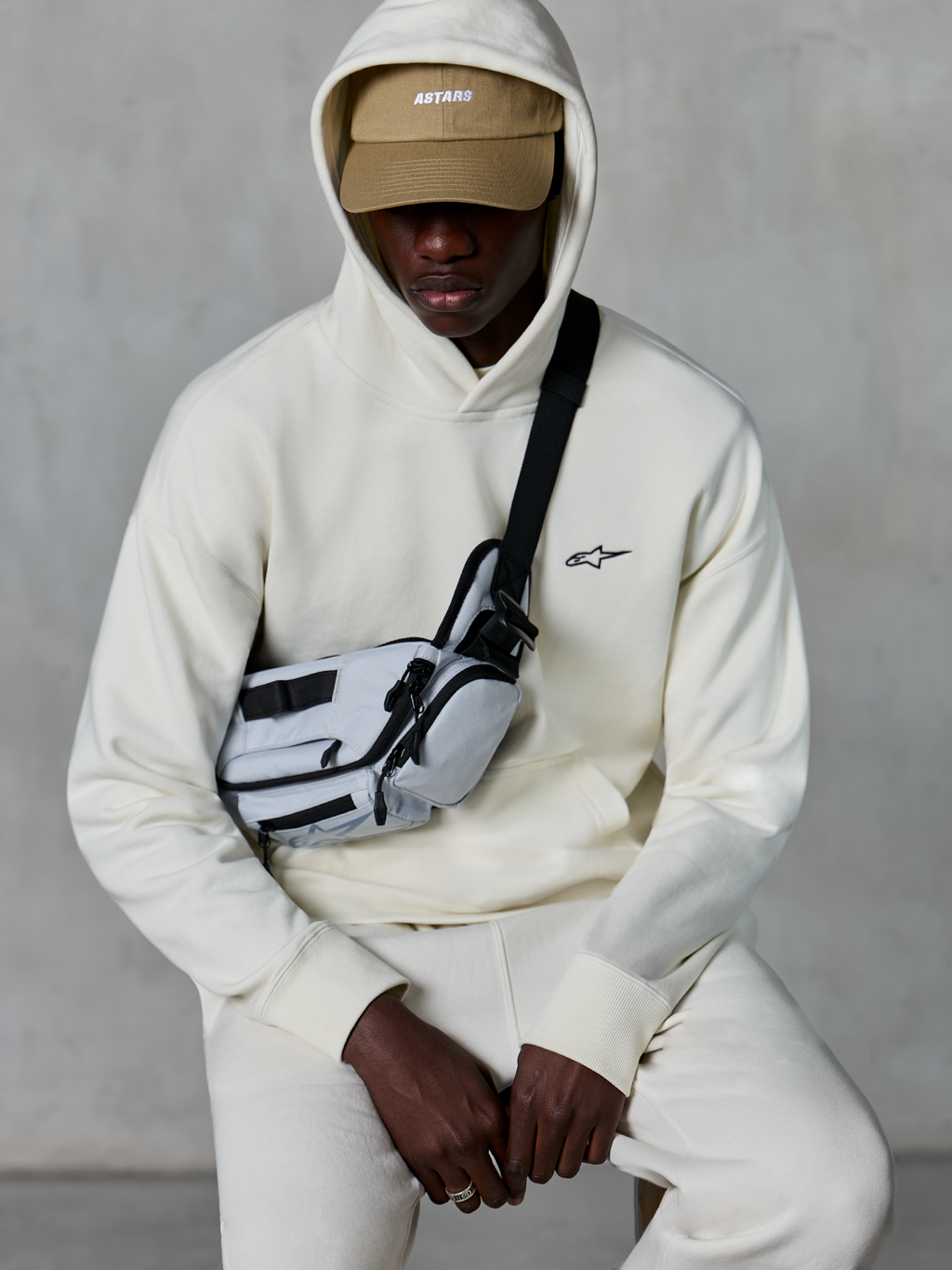 Alpinestars Essentials Ageless Hoodie, lässige Streetwear, Off-White, kleines schwarzes gesticktes Logo auf der Brust, Kapuze über einer beigen Cap mit ASTARS-Branding, kombiniert mit einer hellgrauen Crossbody-Tasche, hochwertige Lifestyle-Bekleidung