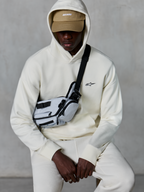Alpinestars Essentials Ageless Hoodie, lässige Streetwear, Off-White, kleines schwarzes gesticktes Logo auf der Brust, Kapuze über einer beigen Cap mit ASTARS-Branding, kombiniert mit einer hellgrauen Crossbody-Tasche, hochwertige Lifestyle-Bekleidung