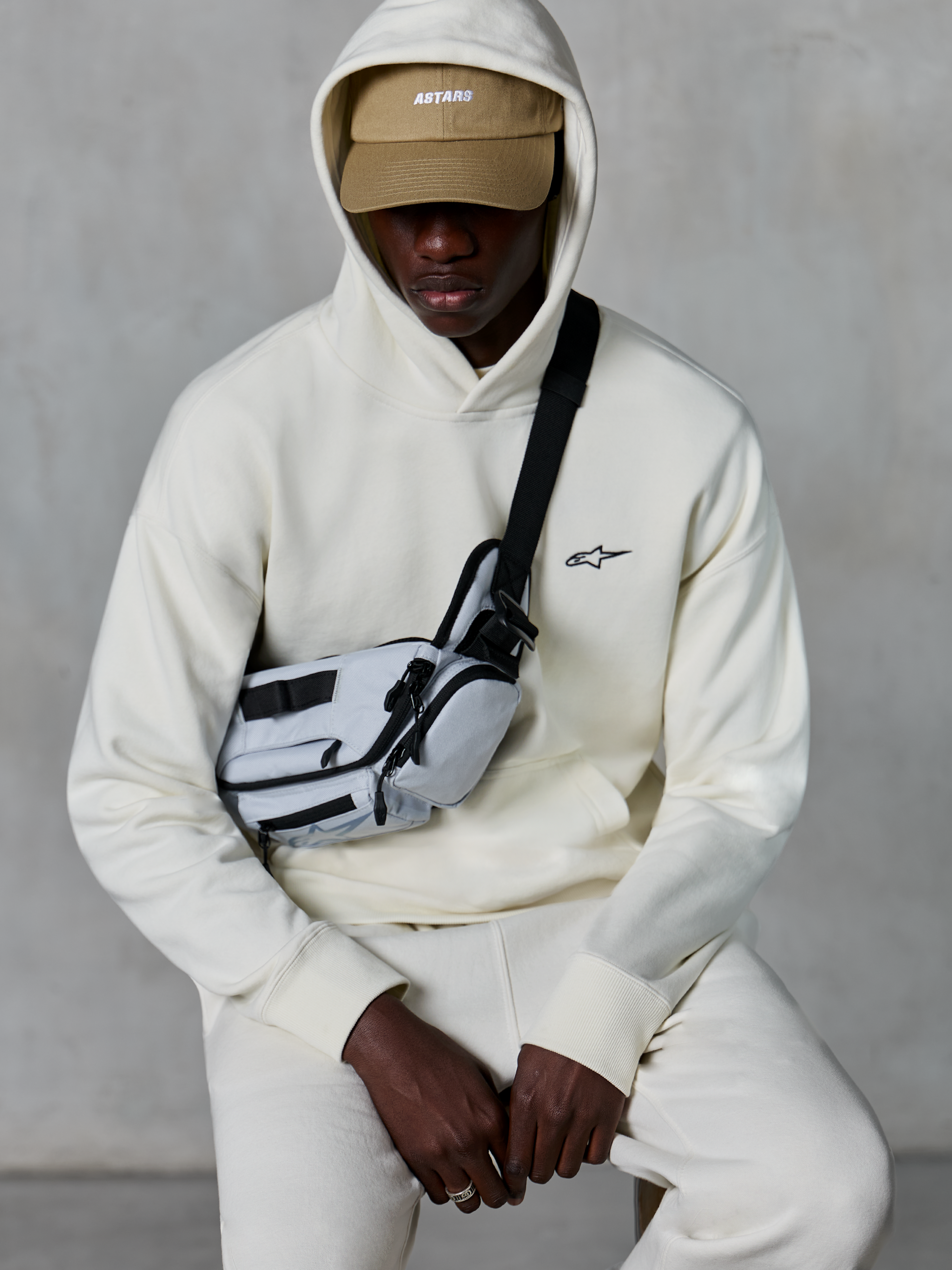 Alpinestars Essentials Ageless Hoodie, lässige Streetwear, Off-White, kleines schwarzes gesticktes Logo auf der Brust, Kapuze über einer beigen Cap mit ASTARS-Branding, kombiniert mit einer hellgrauen Crossbody-Tasche, hochwertige Lifestyle-Bekleidung