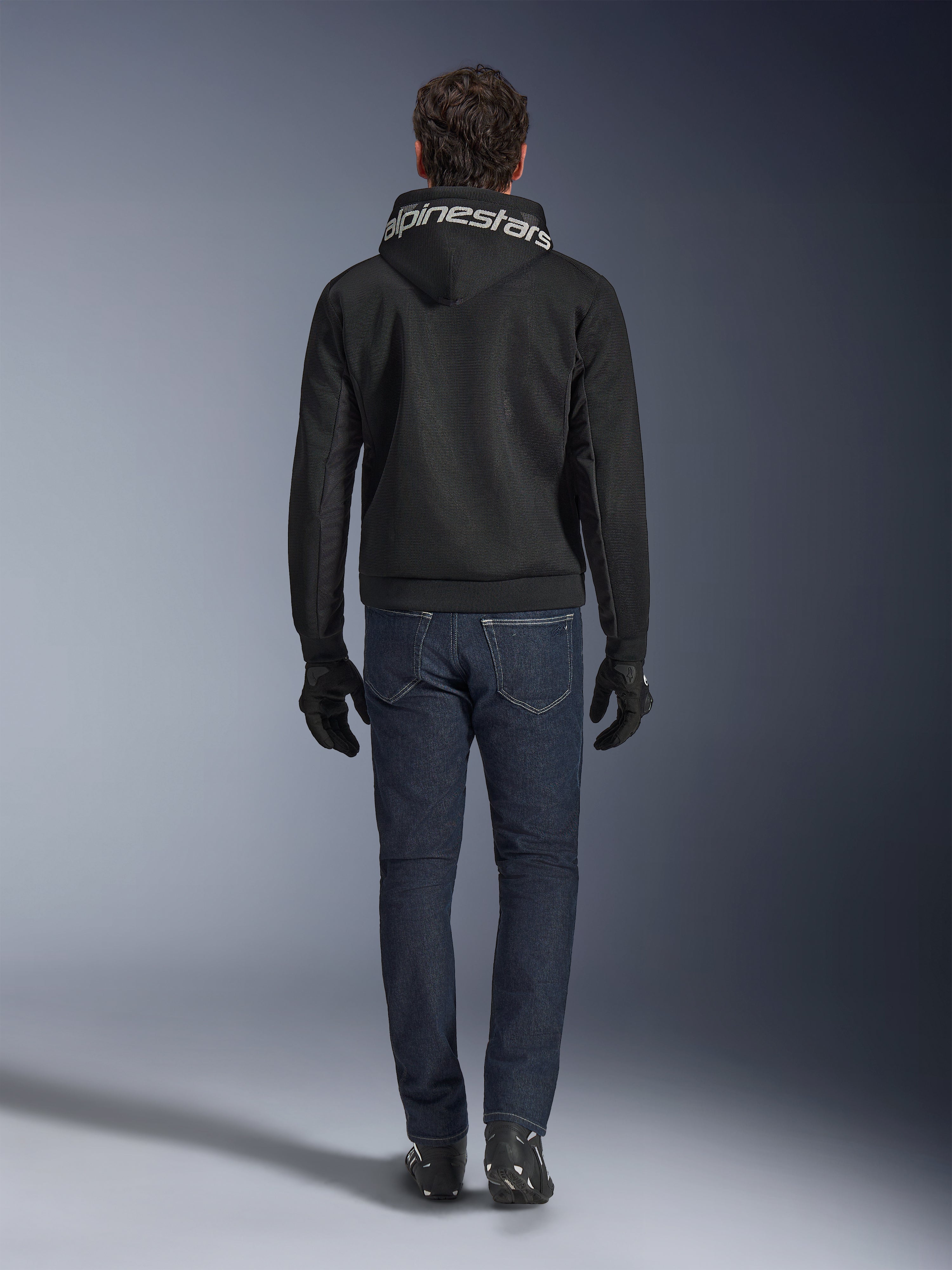 Alpinestars Chrome Superairflow Hoodie, schwarzer Motorrad-Hoodie mit weißem Branding auf der Kapuze, Rückansicht, getragen mit dunkelblauen Jeans, schwarzen Handschuhen und schwarz-weißen Stiefeln, urbane Sport-Fahrausrüstung