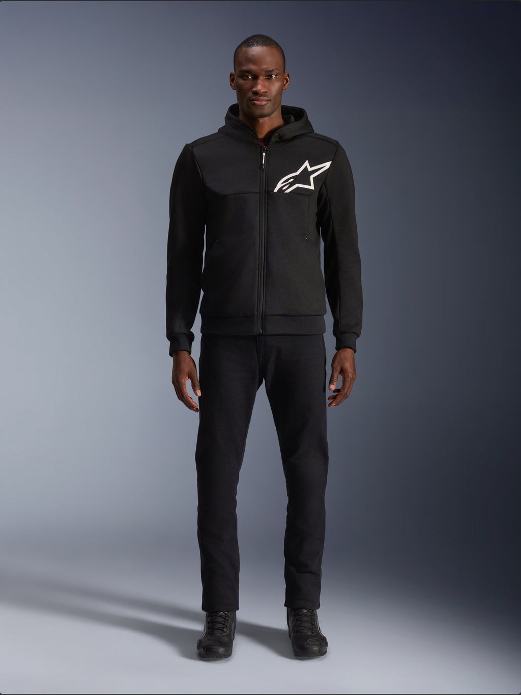 Alpinestars Chrome V2 Sport Hoodie, schwarz-weißer Motorrad-Hoodie, mit einem großen weißen Alpinestars-Logo auf der Brust, Reißverschluss vorne und Kapuze, getragen von einem Modell in schwarzer Hose und Stiefeln, urbane Motorrad-Schutzausrüstung.