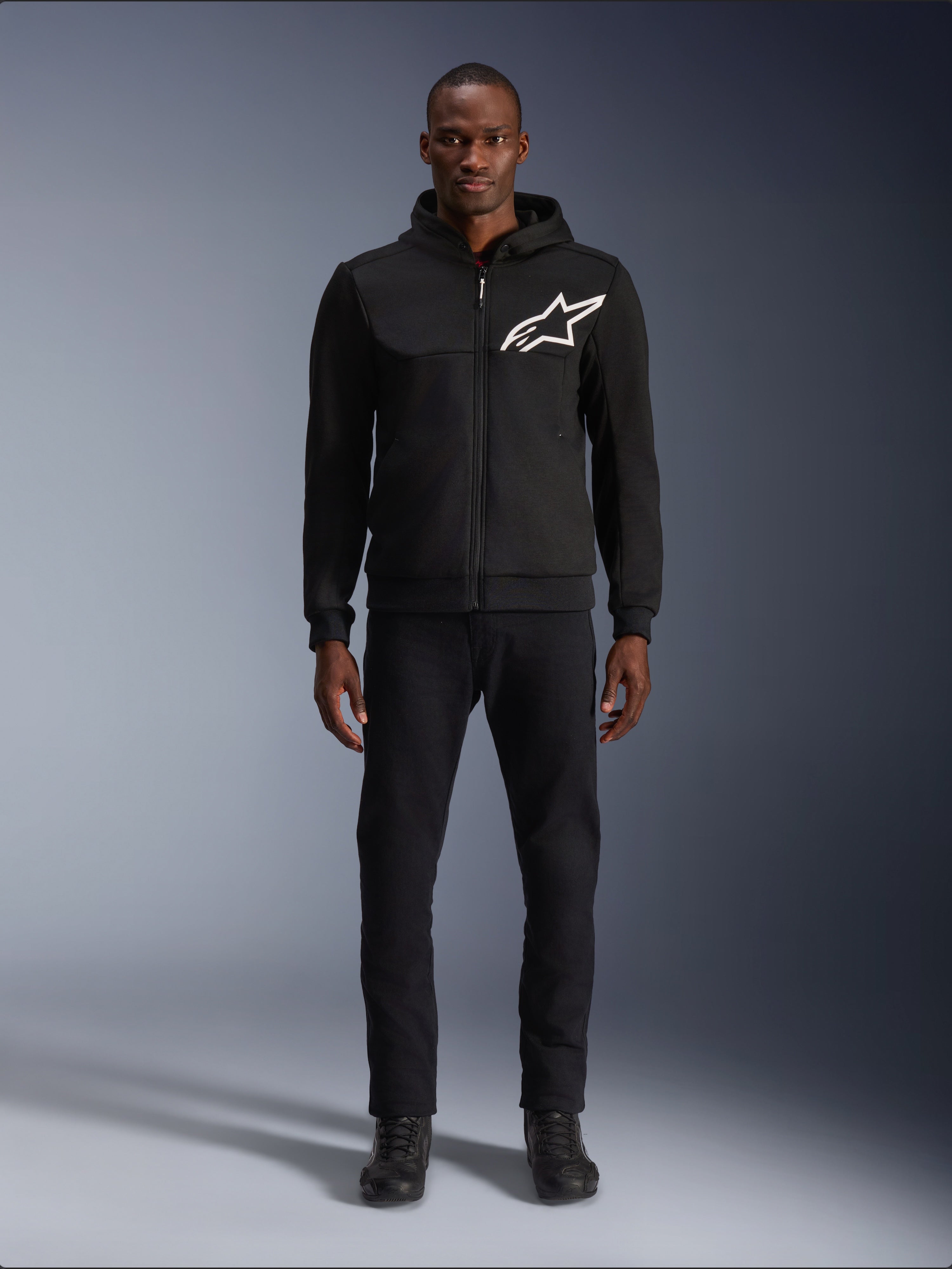 Alpinestars Chrome V2 Sport Hoodie, schwarz-weißer Motorrad-Hoodie, mit einem großen weißen Alpinestars-Logo auf der Brust, Reißverschluss vorne und Kapuze, getragen von einem Modell in schwarzer Hose und Stiefeln, urbane Motorrad-Schutzausrüstung.
