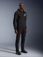 Alpinestars Chrome V2 Sport Hoodie, schwarzer Motorrad-Hoodie mit weißem Stern-Logo, getragen von einem männlichen Model mit passender schwarzer Hose und Motorrad-Sneakern, urbane Lifestyle-Fahrausrüstung