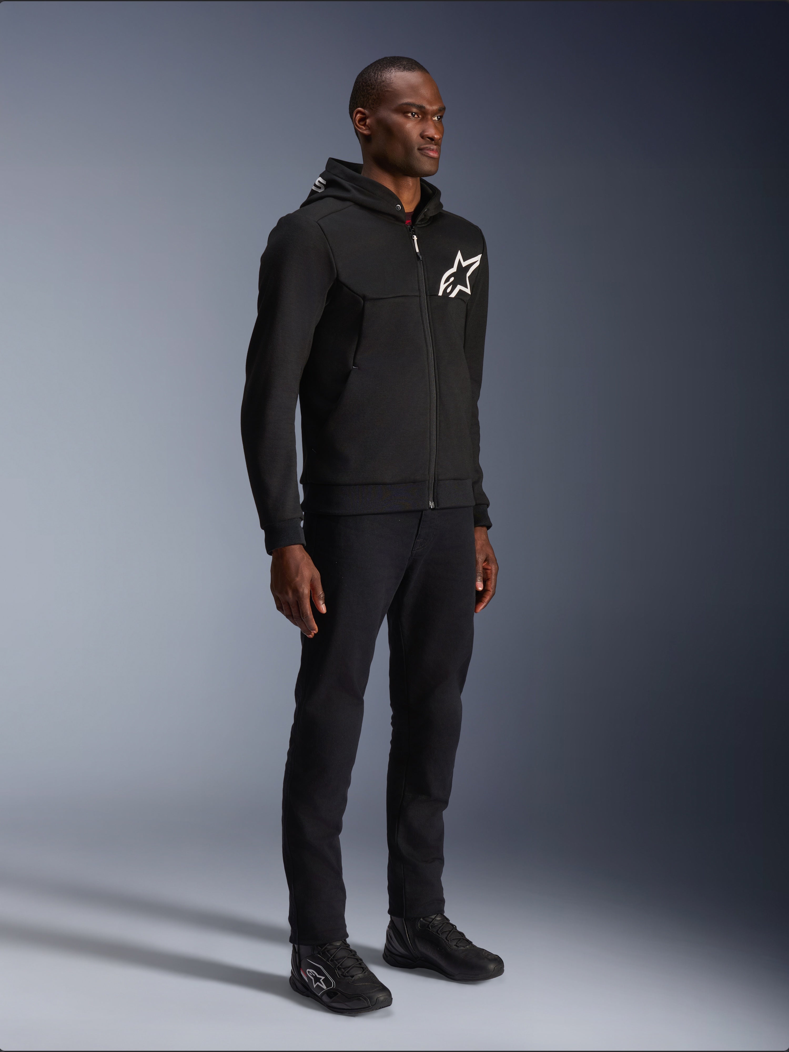 Alpinestars Chrome V2 Sport Hoodie, schwarzer Motorrad-Hoodie mit weißem Stern-Logo, getragen von einem männlichen Model mit passender schwarzer Hose und Motorrad-Sneakern, urbane Lifestyle-Fahrausrüstung