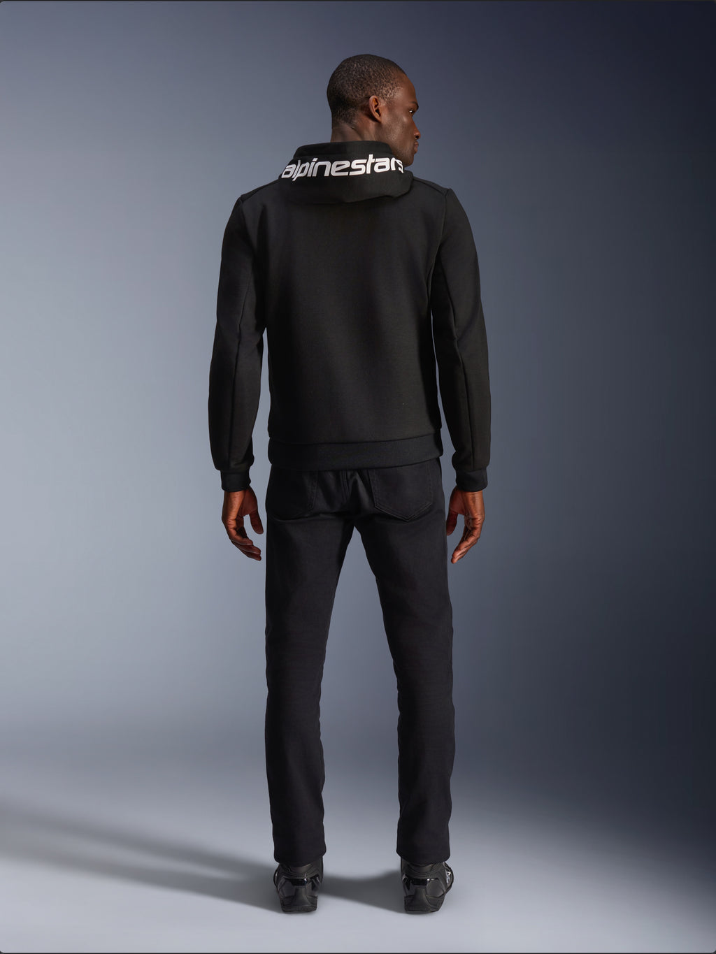 Alpinestars Chrome V2 Sport Hoodie, Motorrad-Hoodie, Schwarz, Rückansicht eines Models mit schwarzem Tech-Fleece-Hoodie und weißem Alpinestars-Logo auf der Kapuze, kombiniert mit schwarzer Hose und Stiefeln, lässiger urbaner Fahrstil