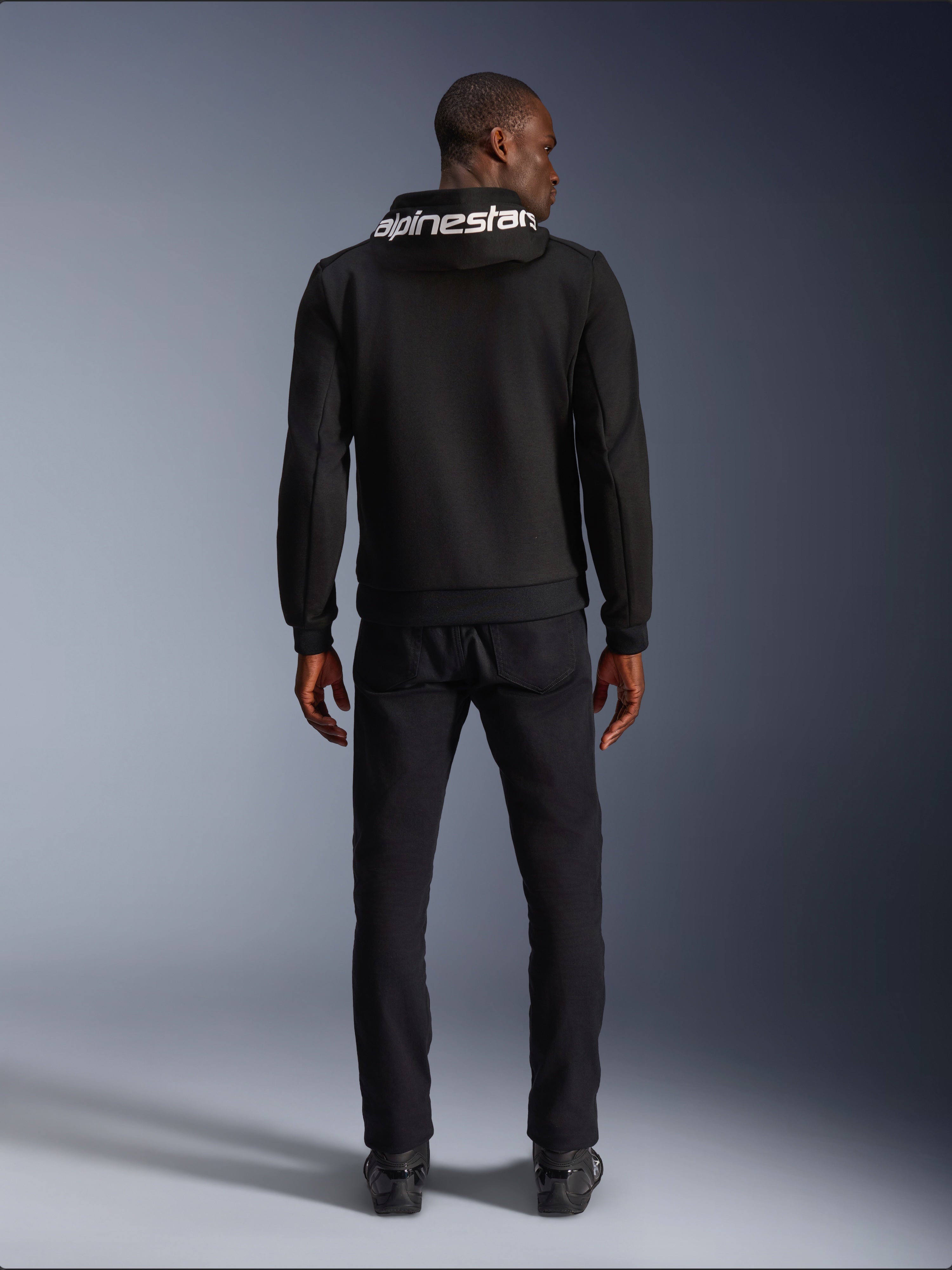 Alpinestars Chrome V2 Sport Hoodie, Motorrad-Hoodie, Schwarz, Rückansicht eines Models mit schwarzem Tech-Fleece-Hoodie und weißem Alpinestars-Logo auf der Kapuze, kombiniert mit schwarzer Hose und Stiefeln, lässiger urbaner Fahrstil