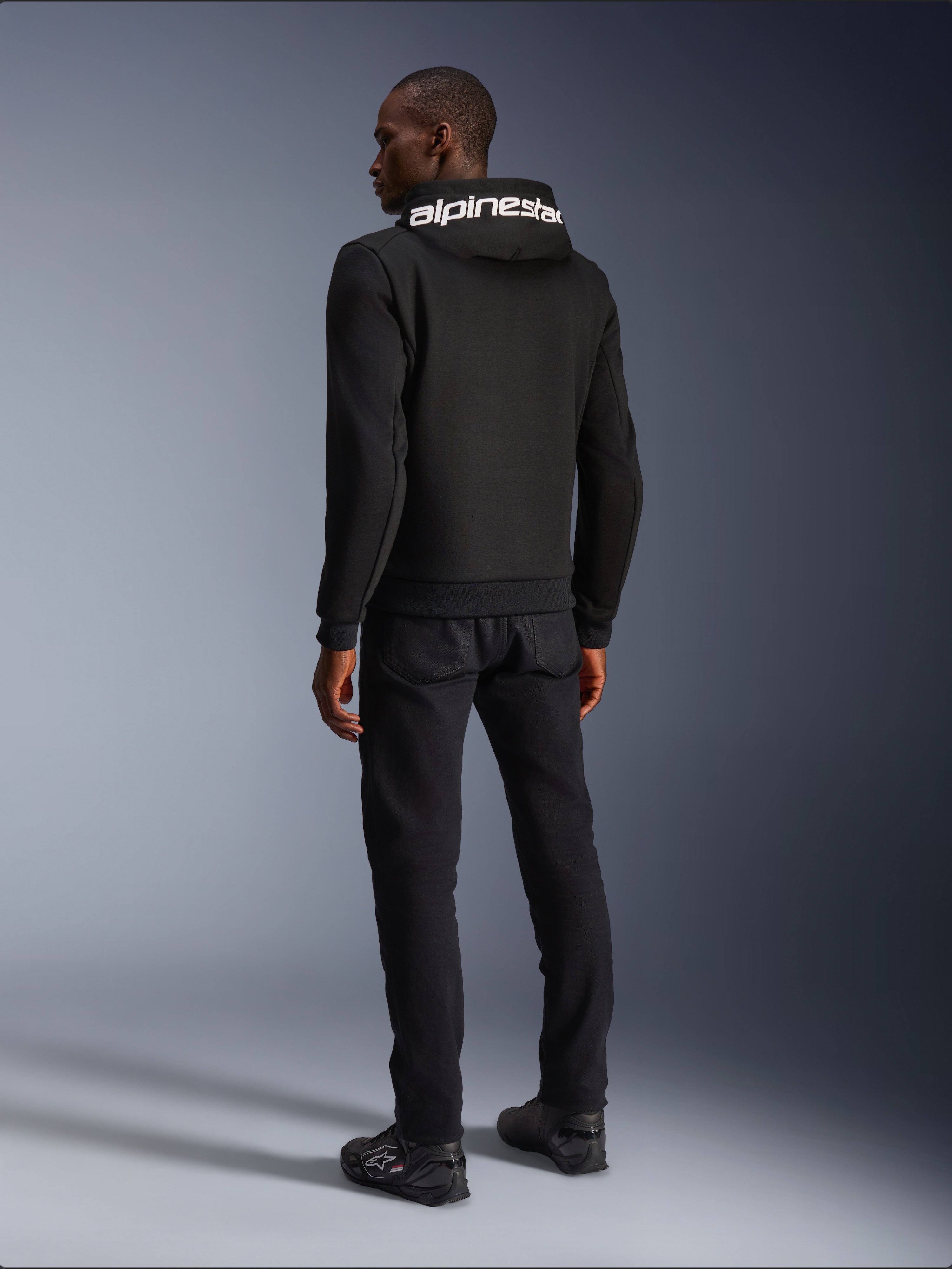 Alpinestars Chrome V2 Sport Hoodie, Schwarz, Rückansicht mit weißem Alpinestars-Branding auf der Kapuze, getragen mit schwarzer Hose und Motorradschuhen, lässige urbane Motorradbekleidung mit athletischer Silhouette