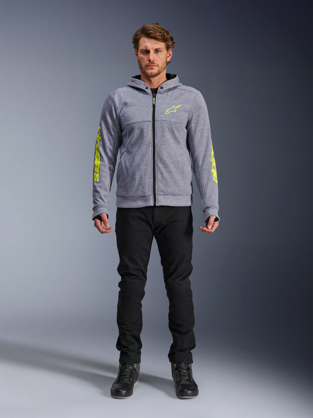 Alpinestars Chrome V2 Sport Hoodie, Grau meliert mit neongelben Akzenten, urbane Motorradbekleidung, kombiniert mit schwarzer Hose und schwarzen Motorradschuhen, technisches Fleece mit auffälligem gelben Branding auf Ärmeln und Brust, lässiger Streetstyle zum Fahren
