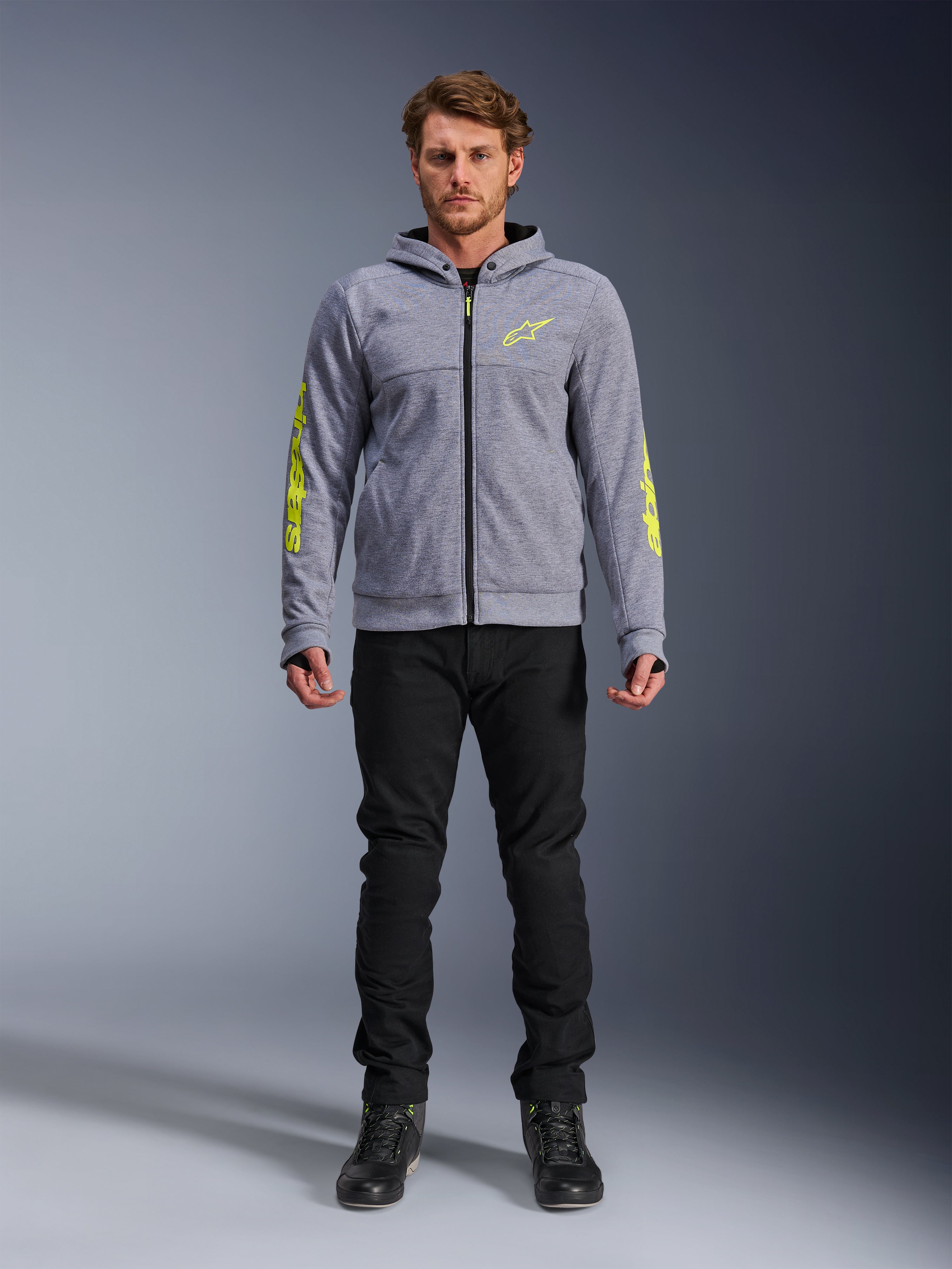 Alpinestars Chrome V2 Sport Hoodie, Grau meliert mit neongelben Akzenten, urbane Motorradbekleidung, kombiniert mit schwarzer Hose und schwarzen Motorradschuhen, technisches Fleece mit auffälligem gelben Branding auf Ärmeln und Brust, lässiger Streetstyle zum Fahren